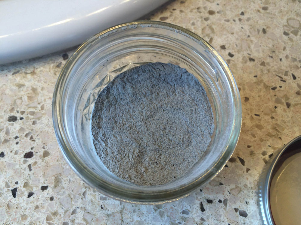 An small mason jar has a gray powder inside of it. This jar sits on a counter top with black, tan, and light colored spots and a white sink.