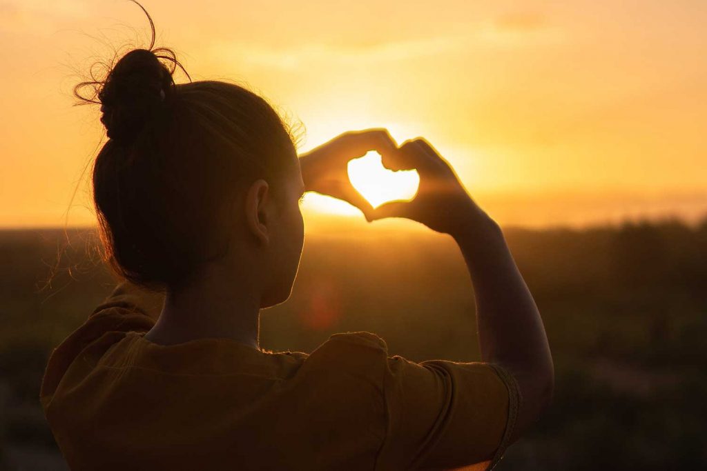 [A girl in a mustard yellow blouse has her hands in a shape of a heart. The sun is shining through the heart from a distance.]