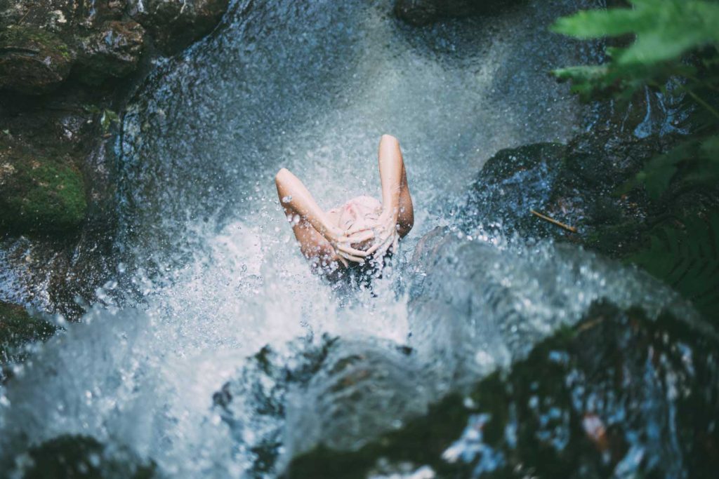 [A light skinned person is showering at the base of a waterfall. The camera is on top of the waterfall.]