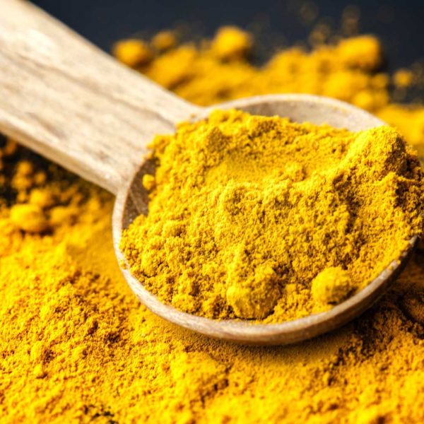 [A light colored wooden spoon has bright gold turmeric in it as well as underneath on a black background.]