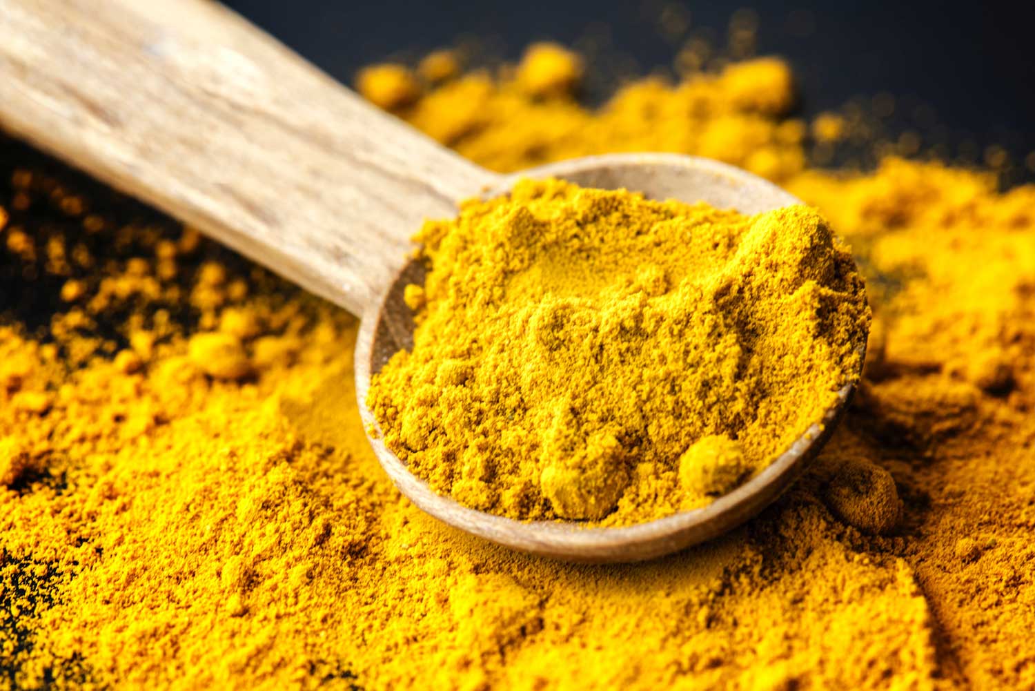 [A light colored wooden spoon has bright gold turmeric in it as well as underneath on a black background.]