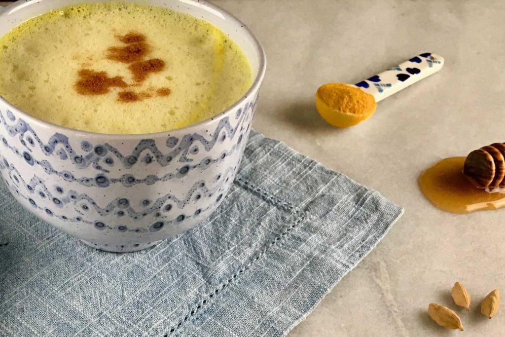 [A white and blue mug has a yellow color tea inside. Underneath the mug is a blue fabric placemat. Next to it is a spoon of turmeric, a stick of honey, and cardamon.]