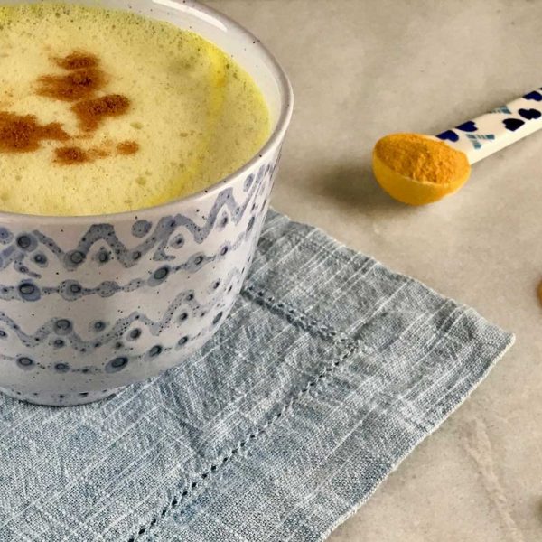 [A white and blue mug has a yellow color tea inside. Underneath the mug is a blue fabric placemat. Next to it is a spoon of turmeric, a stick of honey, and cardamon.]