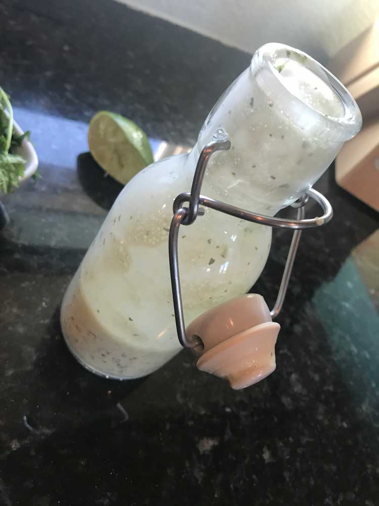 A glass jar with white sauce and green flecks inside is resting on a black granite countertop. A knife block and half of a lime can be see in the blurry background.