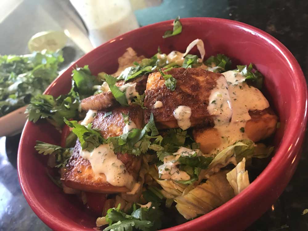 A red bowl contains cilantro, lettuce, and tofu drizzled with a white-green sauce. In the background, a bowl of cilantro, half of a lime, and a glass of the sauce is blurred.