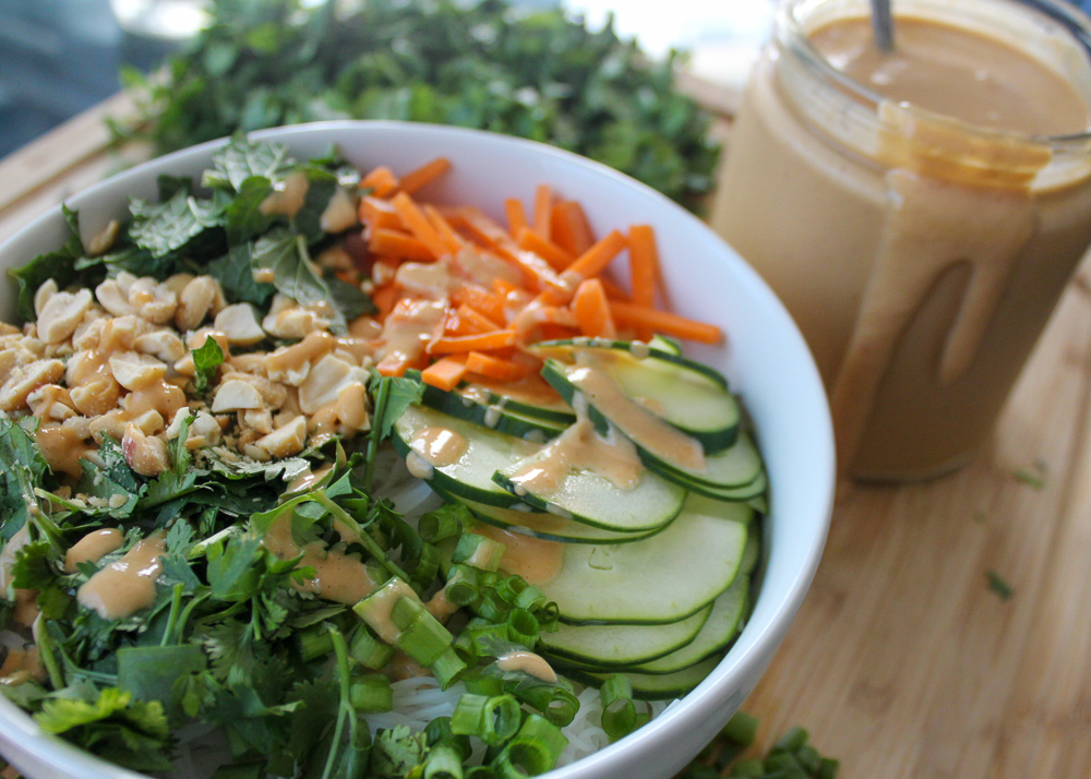 A white bowl holding white noodles is topped with sliced cucumber, julienned carrots, crushed peanuts, chopped cilantro, mint, and scallions. Next to it is a jar with brown sauce and a pile of greens.