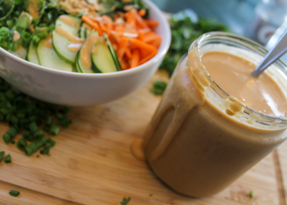 A jar of brown sauce is on a wooden cutting board. Next to the jar is a blurred pile of greens and a white bowl with colorful vegetables. The rest of the background is blurred.