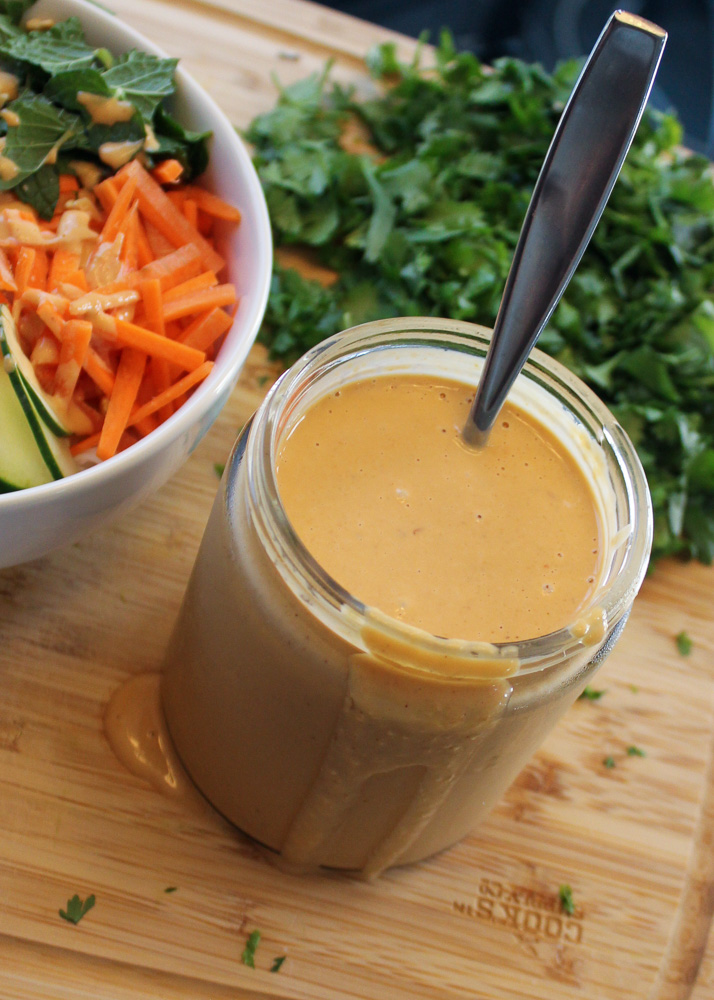 A jar of brown sauce is on a wooden cutting board. Next to the jar is a blurred pile of greens and a white bowl with carrots, cucumber, and mint leaves.