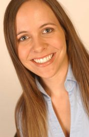 A light skinned person is smiling at the camera. Her brown hair is smooth and long. She is wearing a blue button up shirt.