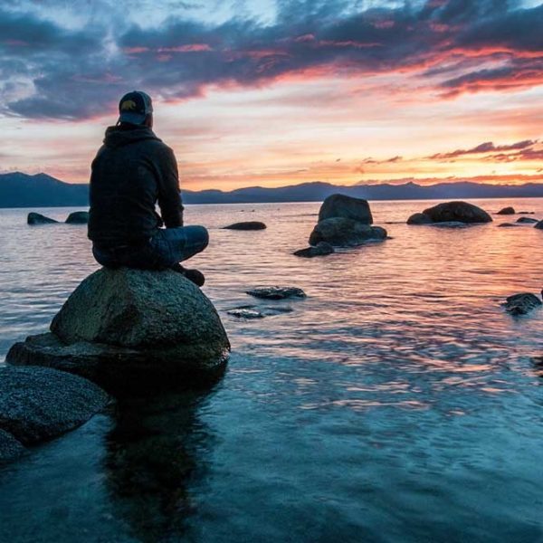 A masculine figure is sitting on a boulder surrounded by semi-calm water. In the distance is the silhouette of mountains lit by the sunset behind it.
