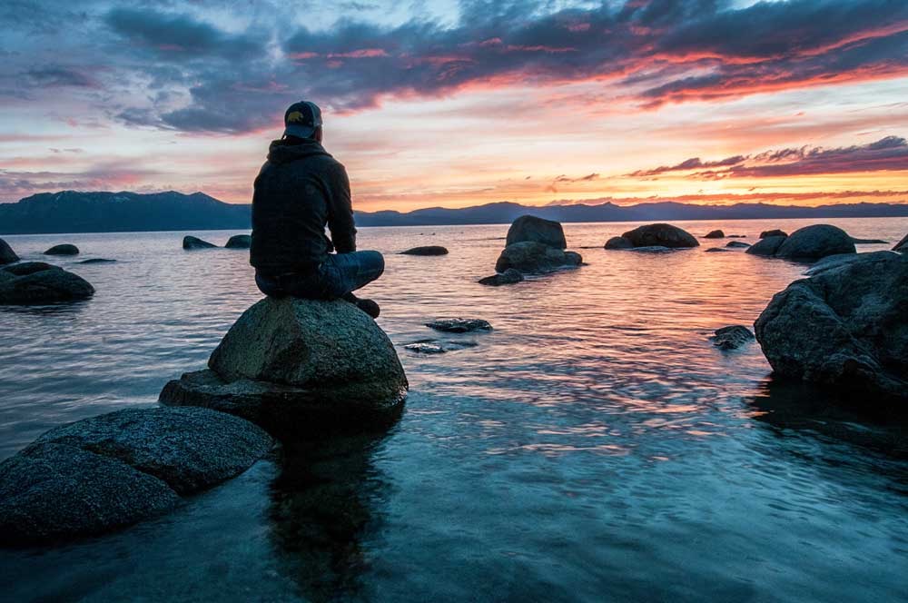 A masculine figure is sitting on a boulder surrounded by semi-calm water. In the distance is the silhouette of mountains lit by the sunset behind it.