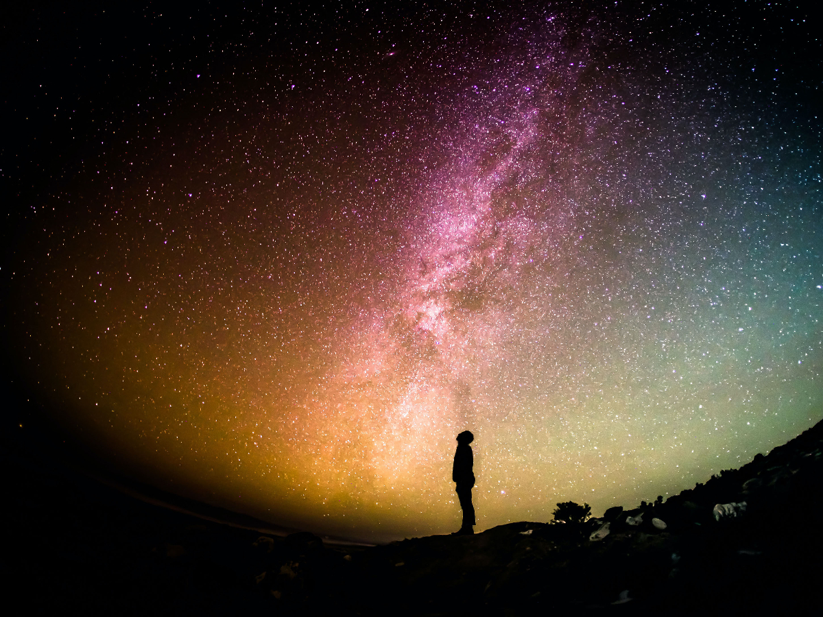 A silhouette of a person is looking up at the night sky. The sky is a dark rainbow ombre with many speckles of stars.