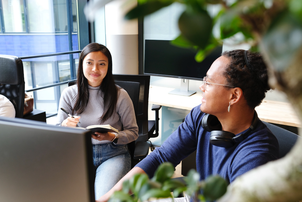 Two persons are happily sitting in a meeting within a modern office with plants. One person is wearing a gray long sleeve and writing in a black journal in the air while the other person wears a blue shirt and large headphones.